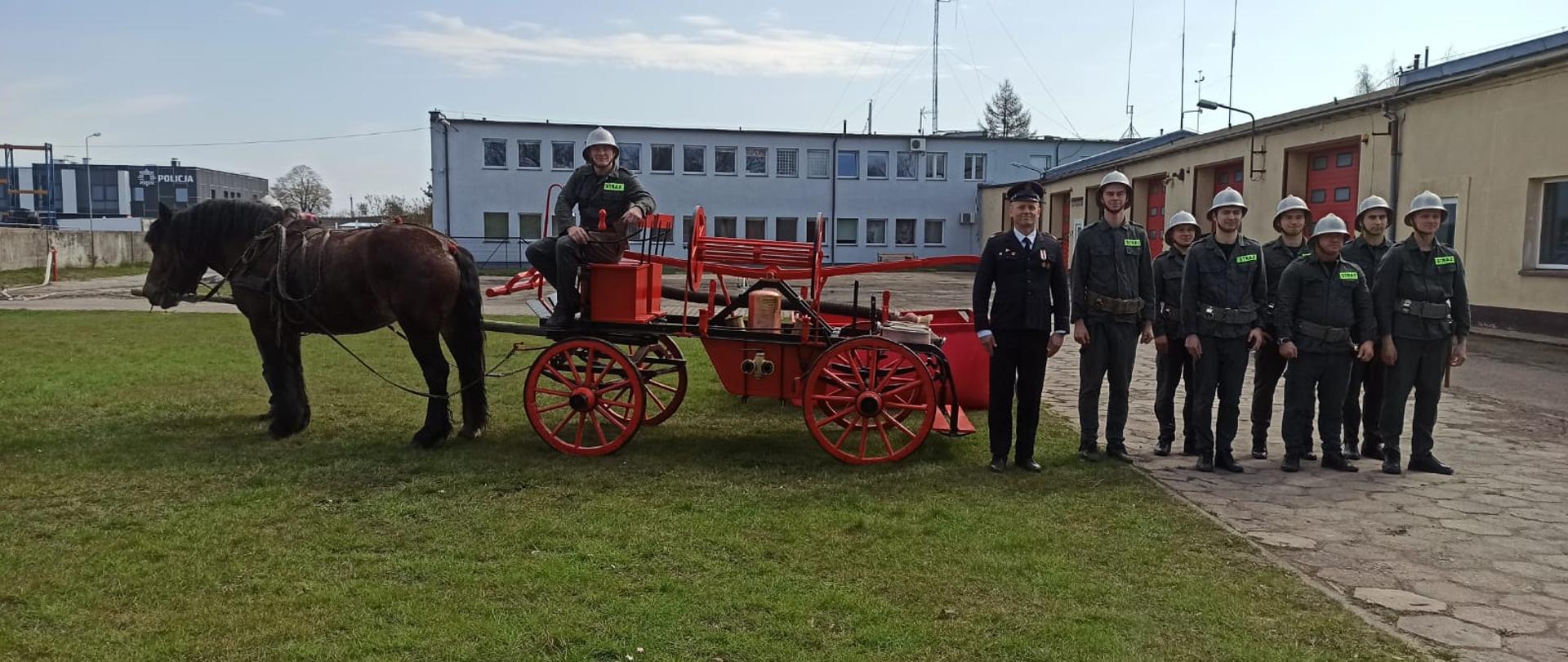 Zdjęcie przedstawia sikawkę konną wraz z drużyną na tle budynków KP PSP Świdwin
