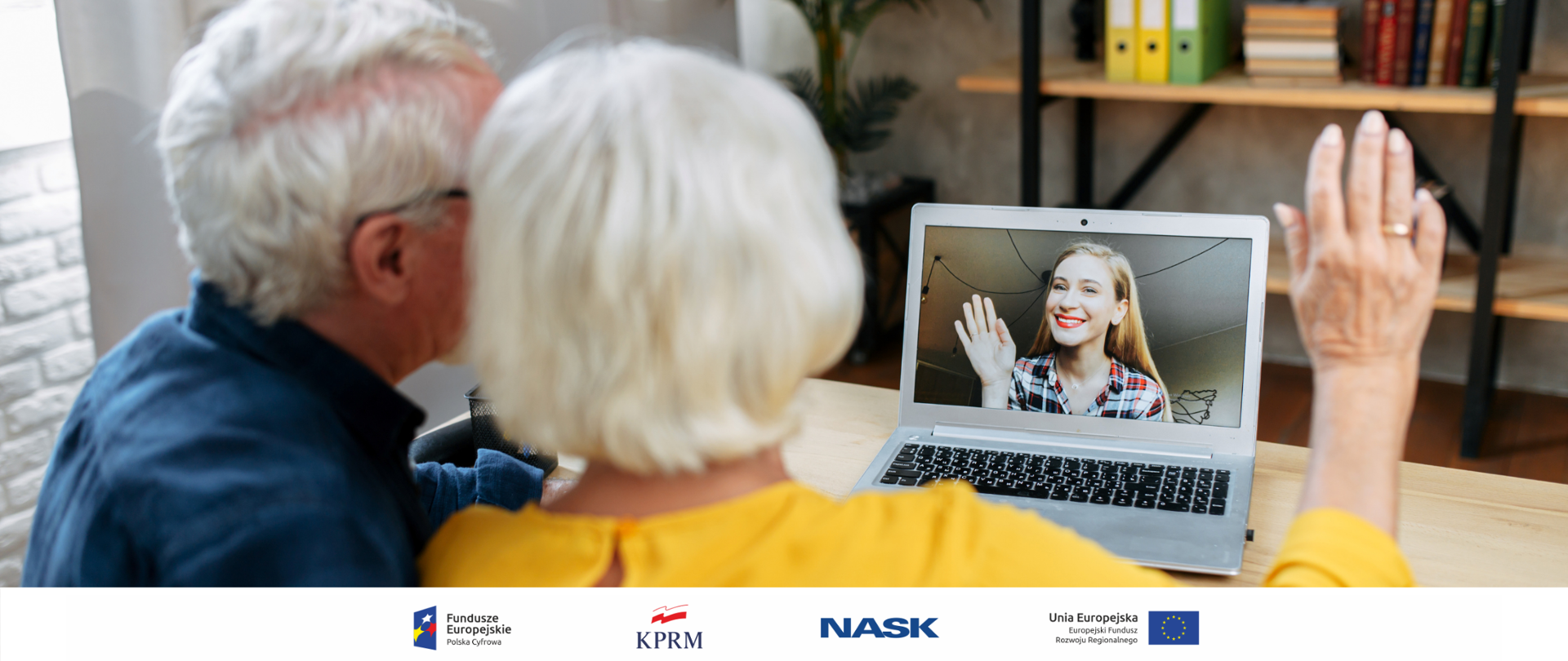 Dwoje seniorów przed laptopem, w trakcie videorozmowy - widok od tyłu. Na ekranie laptopa widoczna młoda, machająca im kobieta (wnuczka).