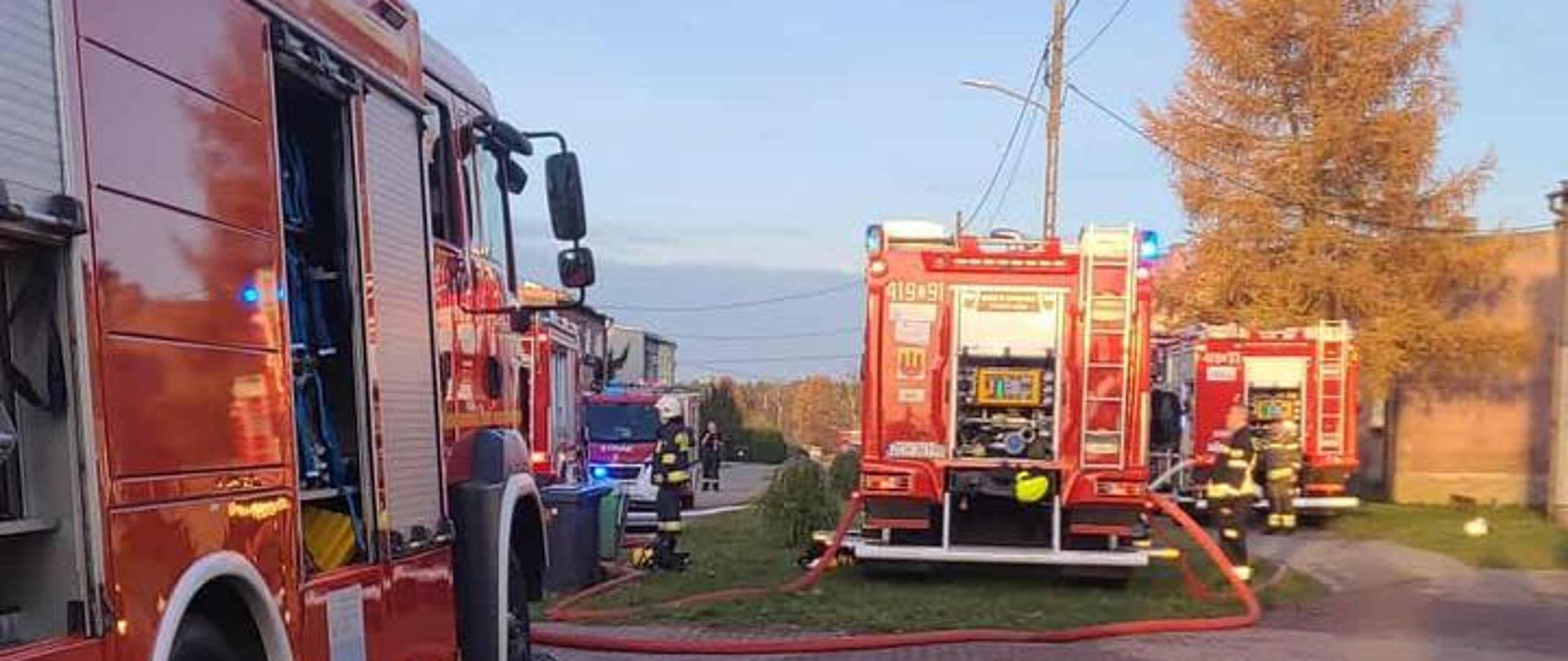 Kilka wozów strażackich ustawionych wzdłuż drogi podczas akcji gaśniczej. Strażacy pracują przy pojazdach, a z węży strażackich prowadzone są linie wodne.
