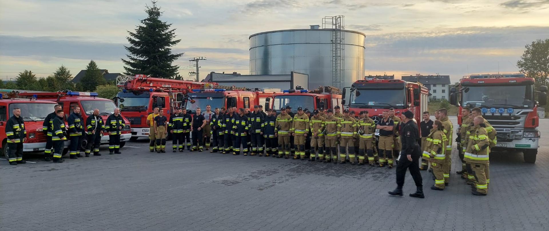 Na brukowanym placu w dwurzędzie stoi grupa strażaków, odbywa się odprawa. Za nimi ustawione jest 7 wozów strażackich. Na końcu placu znajduje się nadziemny zbiornik do celów przeciwpożarowych.