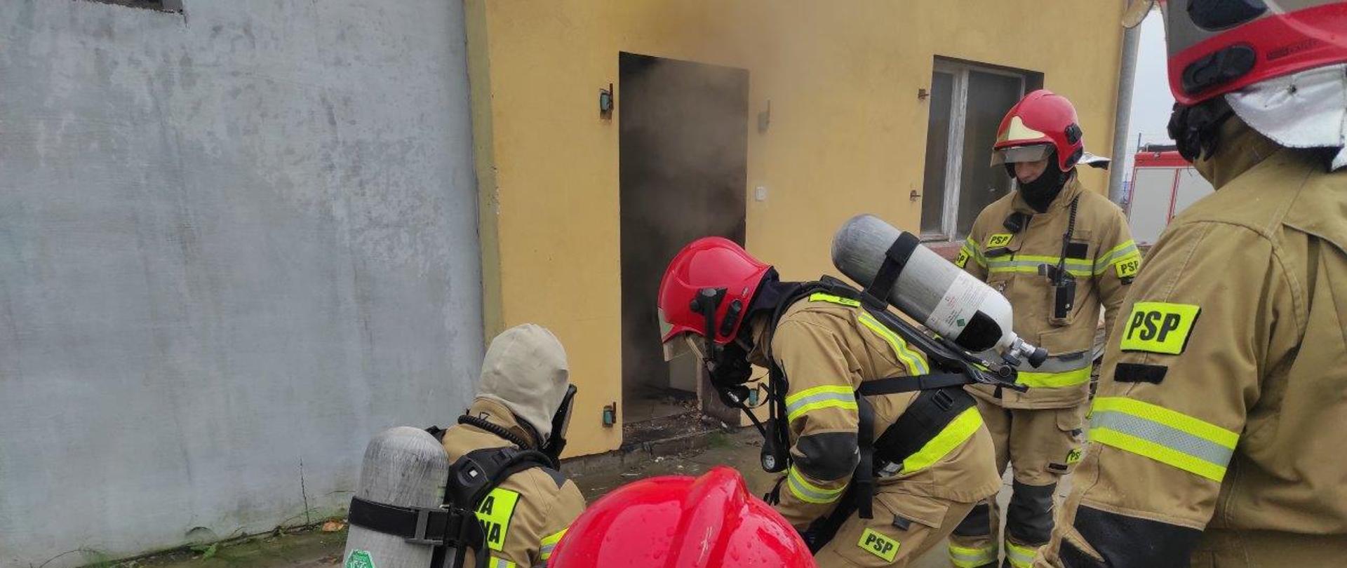 Zdjęcie przedstawia pięciu strażaków ubranych w ubrania ochronne w kolorze piaskowym w hełmach szykujących się do wejścia do wnętrza zadymionego budynku. 