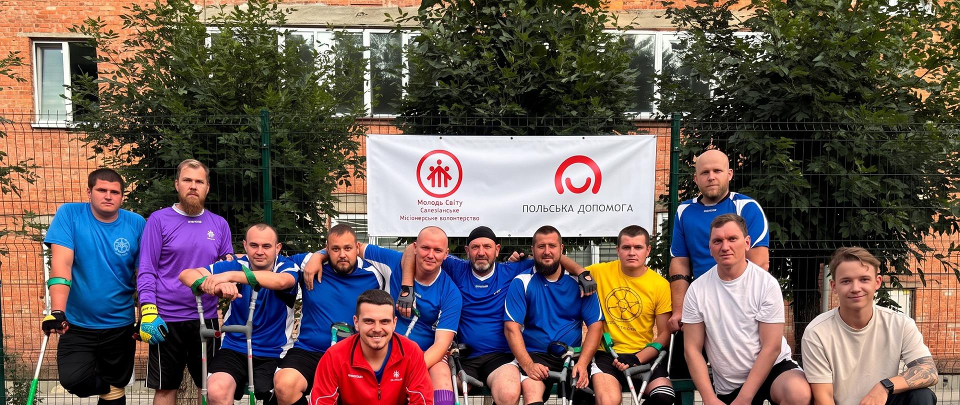 Grupa trzynastu osób pozuje do wspólnego zdjęcia na boisku sportowym. Część osób ma na sobie niebieskie stroje piłkarskie i korzysta z kul łokciowych. Kilka osób ma widoczne amputacje nóg. Za grupą znajduje się baner ze znakami Salezjańskiego Wolontariatu Misyjnego „Młodzi Światu” i programu Polska pomoc oraz napisami w języku ukraińskim. W tle widać ogrodzenie, drzewa i ceglany budynek.
