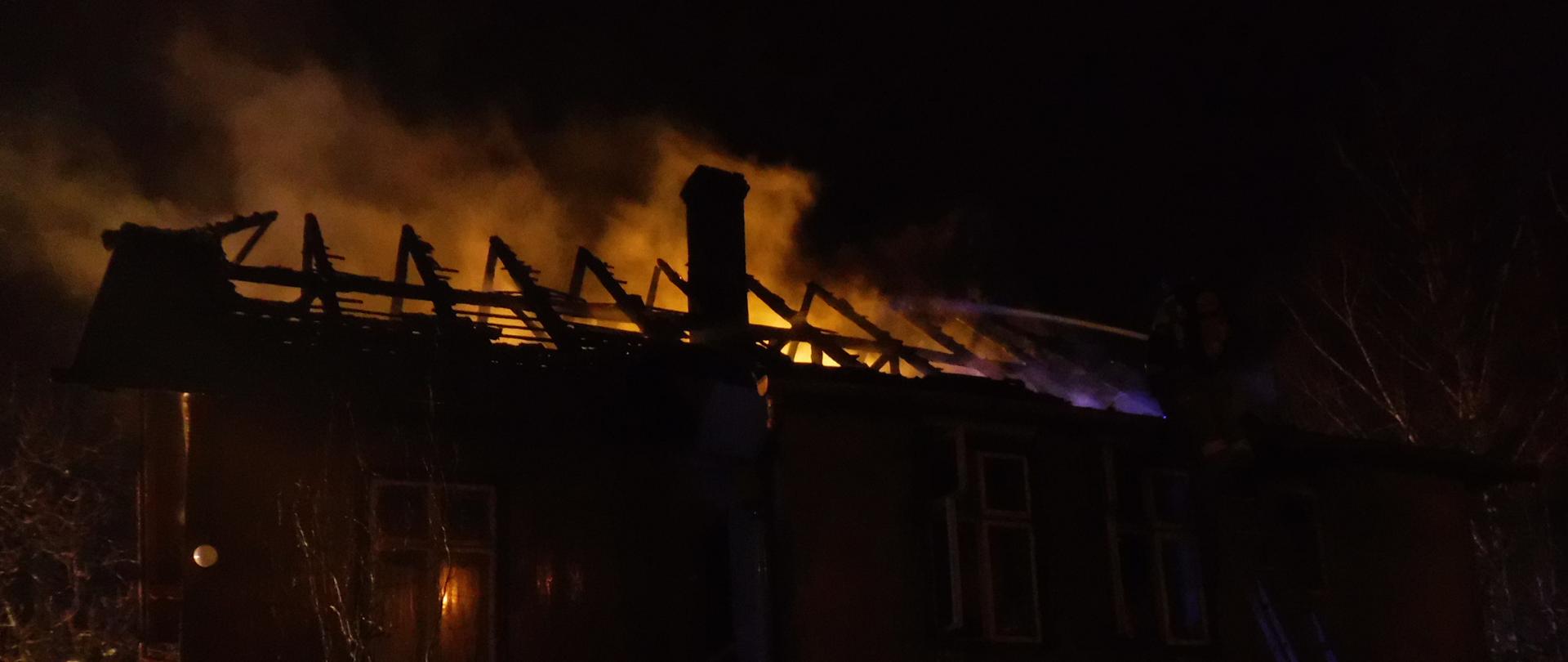 Drewniany budynek mieszkalny, w którym miał miejsce pożar. Na zdjęciu po prawej stronie możemy zaobserwować dwóch strażaków. Jeden z nich przytrzymuje drabinę, na której jest umiejscowiony drugi strażak wraz z jednym prądem wody. Trwa dogaszanie pozostałych po pożarze elementów wierzby dachowej. Nad budynkiem unosi się para wodna wraz z dymem powstałe w wyniku polewania spalonych elementów dachu. W konturach zdjęcia znacząco możemy zaobserwować komin budynku.