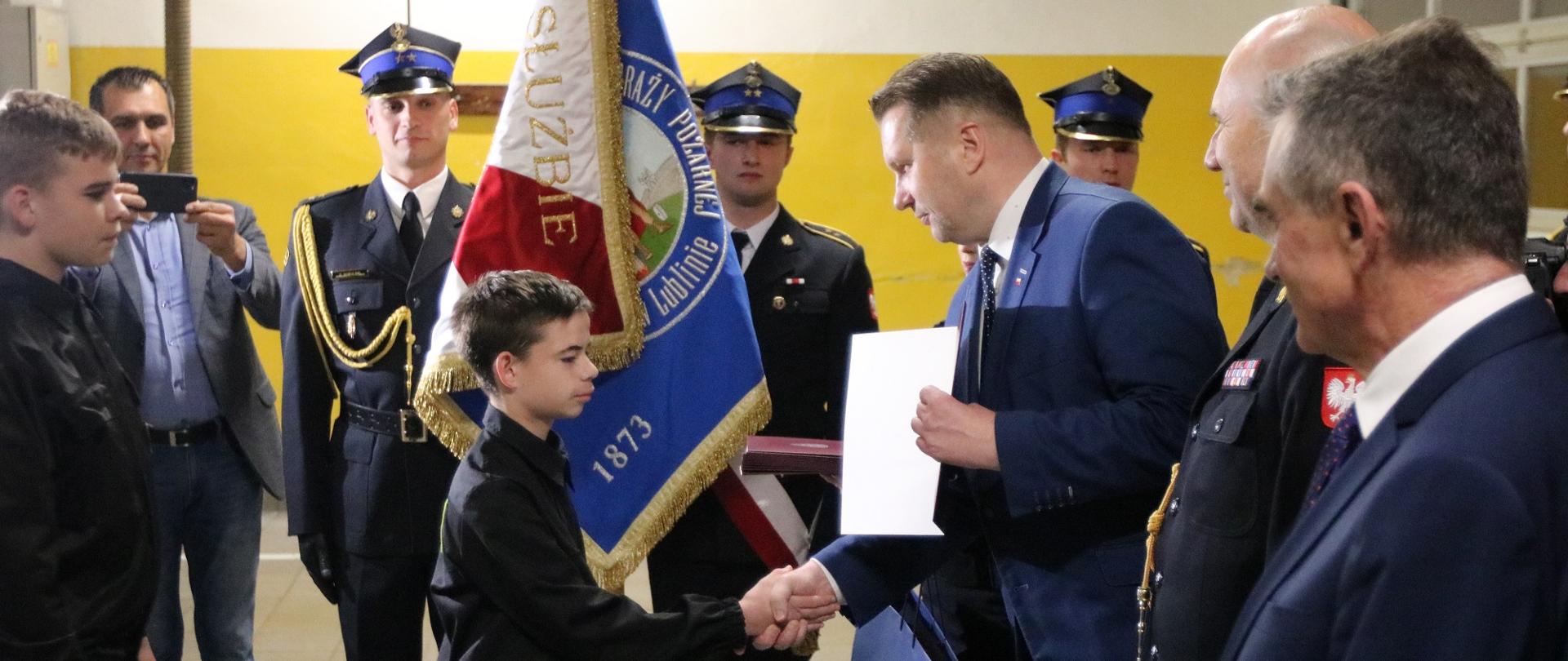 Minister Czarnek ściska rękę małemu chłopcu w czarnym ubraniu, za nimi stoi poczet sztandarowy.