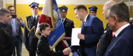 Minister Czarnek ściska rękę małemu chłopcu w czarnym ubraniu, za nimi stoi poczet sztandarowy.