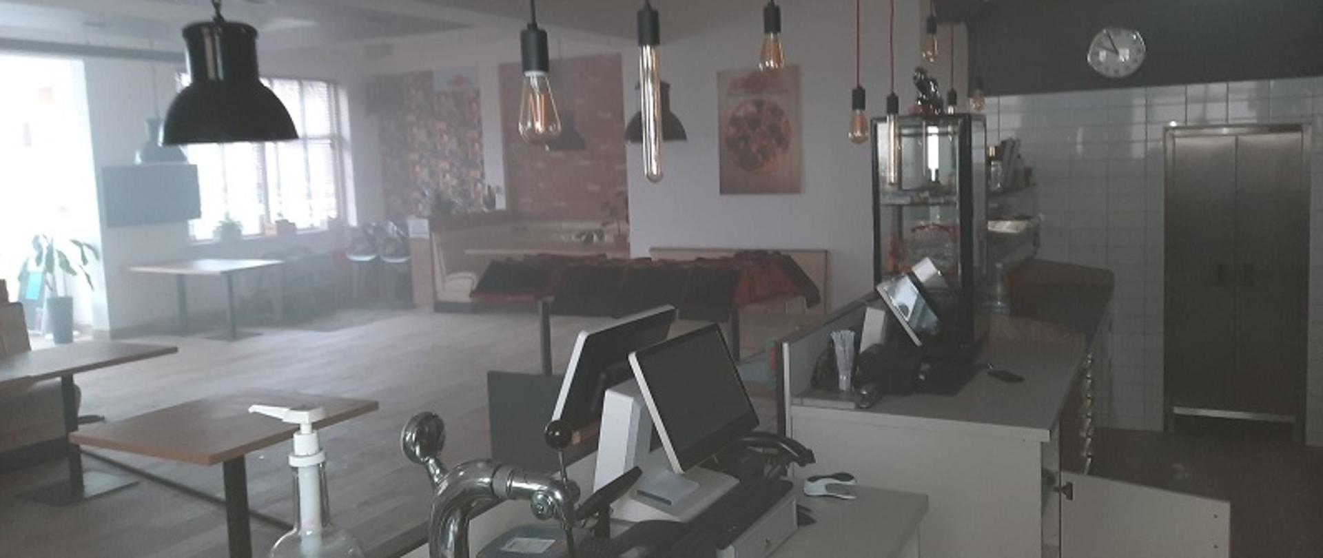 Zdjęcie przedstawia zadymione pomieszczenie lokalu gastronomicznego od strony bufetu. W głębi widoczne stoły dla klientów, ściany ozdobione obrazami i makatami