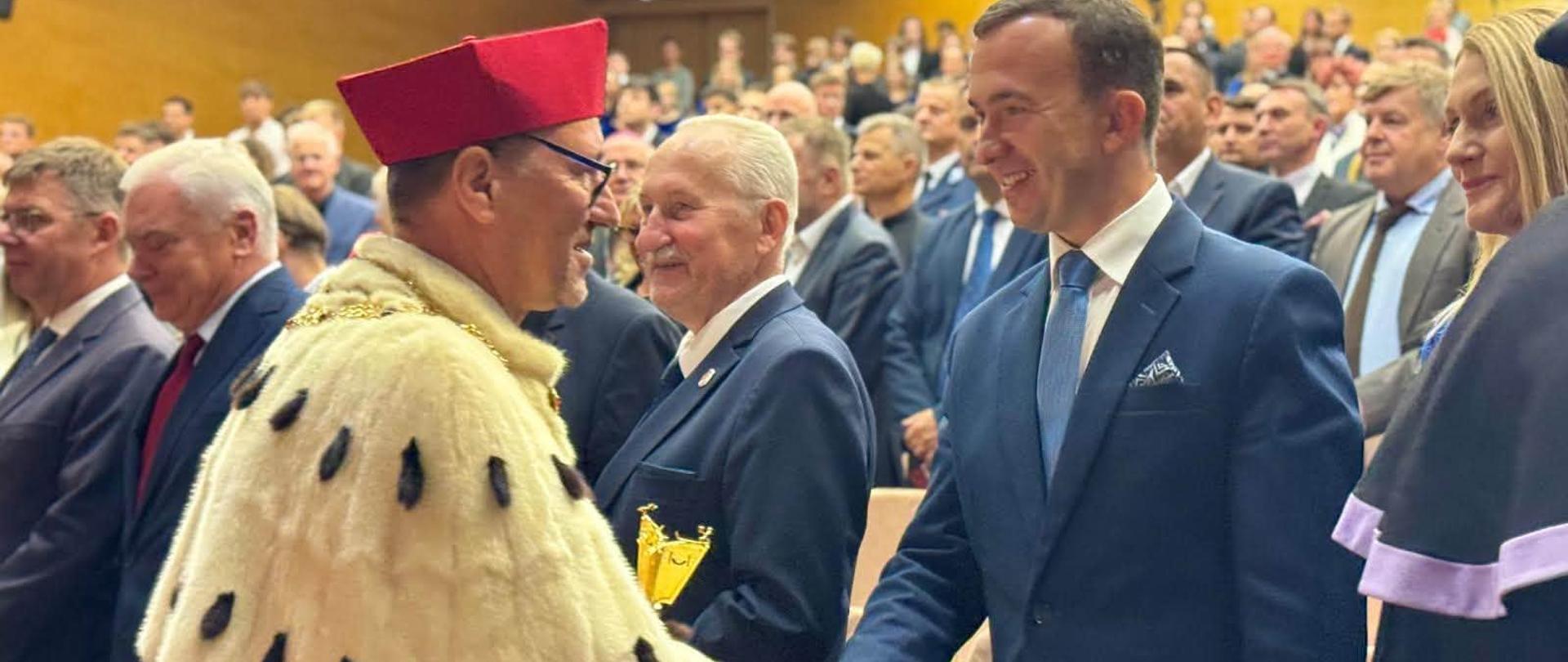 Wicewojewoda w granatowym garniturze podaje rękę rektorowi uczelni w uroczystym stroju akademickim.
