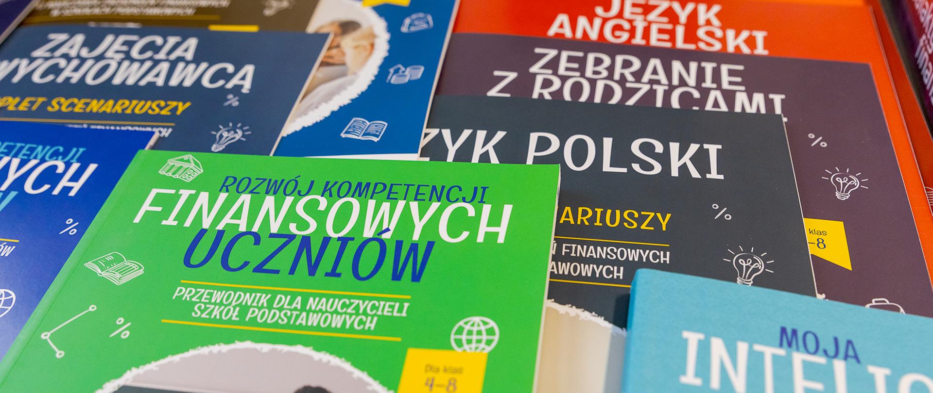Na zdjęciu widoczne są kolorowe publikacje edukacyjne ułożone częściowo jedna na drugiej. Na pierwszym planie znajduje się podręcznik „Rozwój kompetencji finansowych uczniów”, a w tle widoczne są materiały dotyczące języka polskiego, języka angielskiego i zajęć wychowawczych. Okładki zawierają elementy graficzne związane z nauką i edukacją.