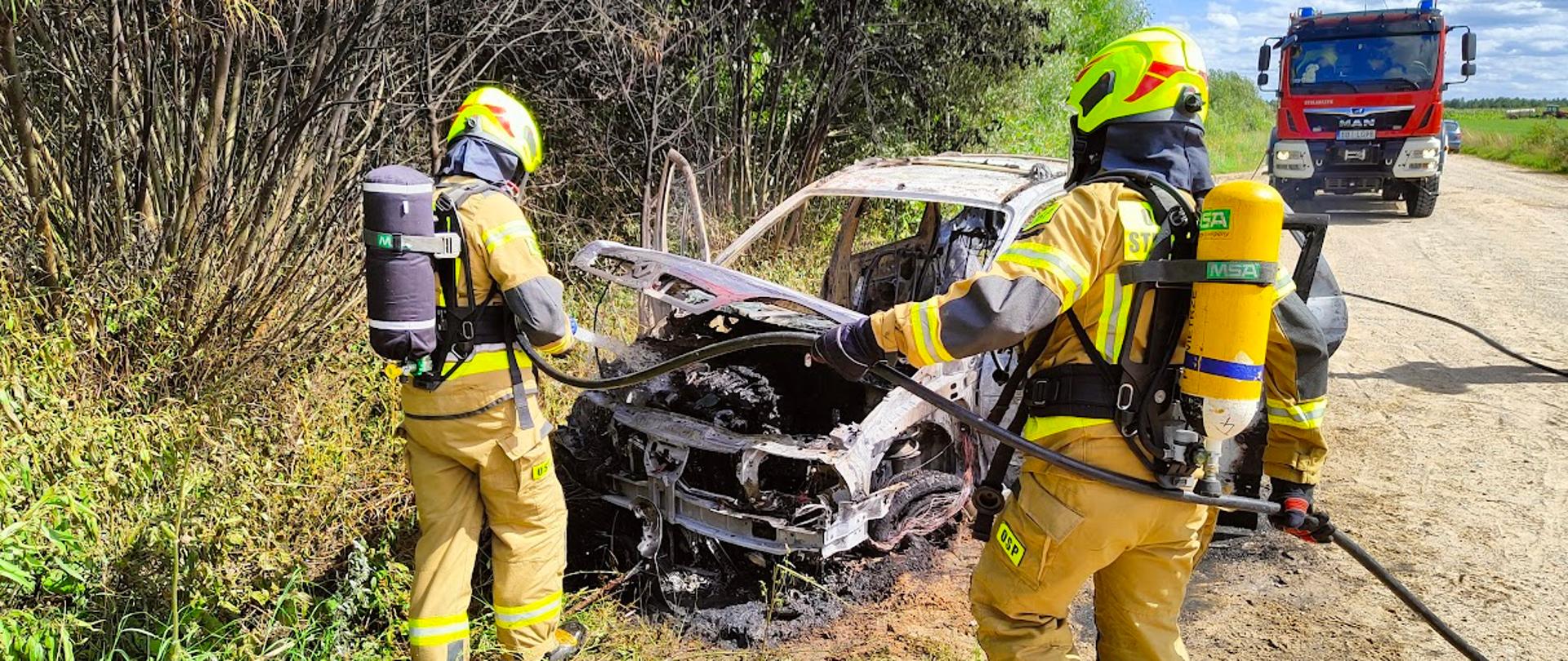Zdjęcie przedstawia dwóch strażaków w ubraniach specjalnych gaszących samochód osobowy na poboczu szutrowej drogi. W tle widać samochód ratowniczo-gaśniczy straży pożarnej.