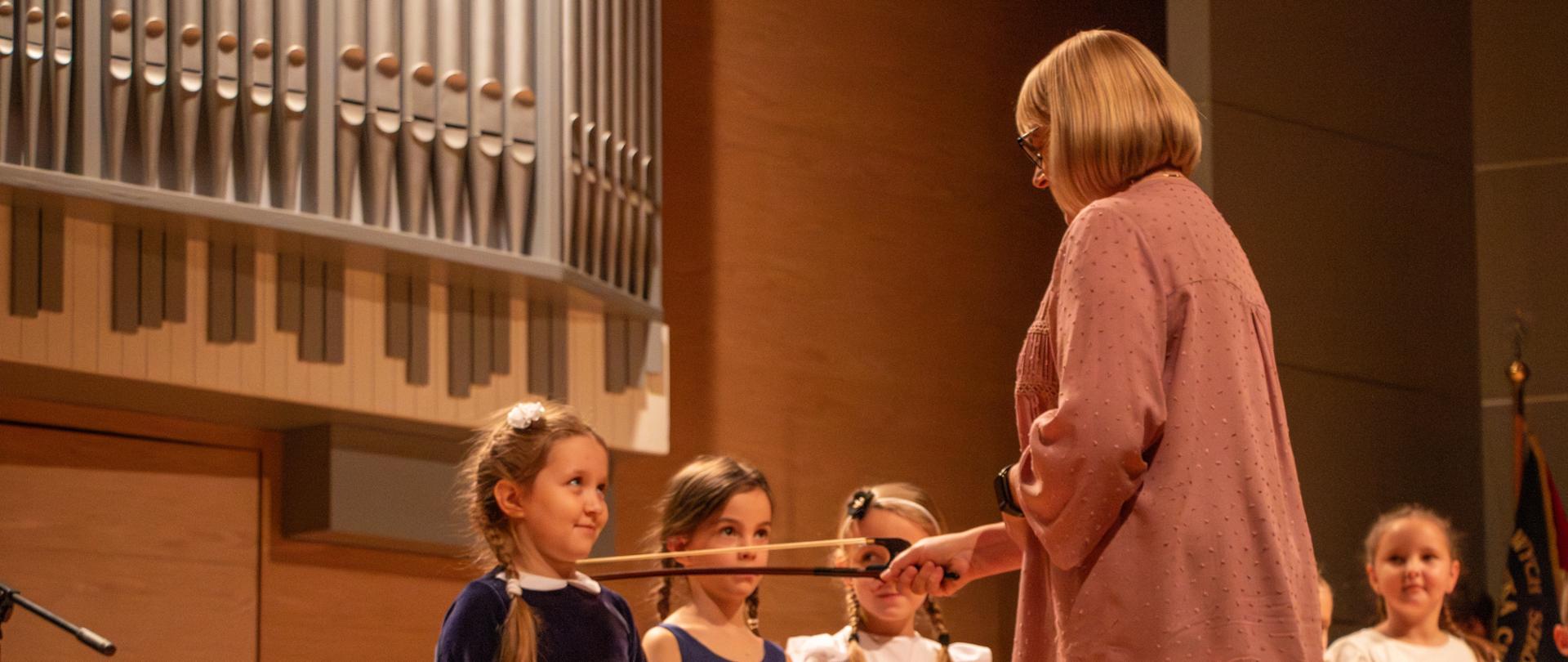 Nauczycielka w różowej bluzce, stojąca z prawej strony, dotyka ramieniem małej dziewczynki ubranej w granatową sukienkę, trzymając w dłoni patyczek symbolizujący pasowanie. Obok niej stoją inne dzieci w odświętnych strojach, a w tle widać część organów piszczałkowych.