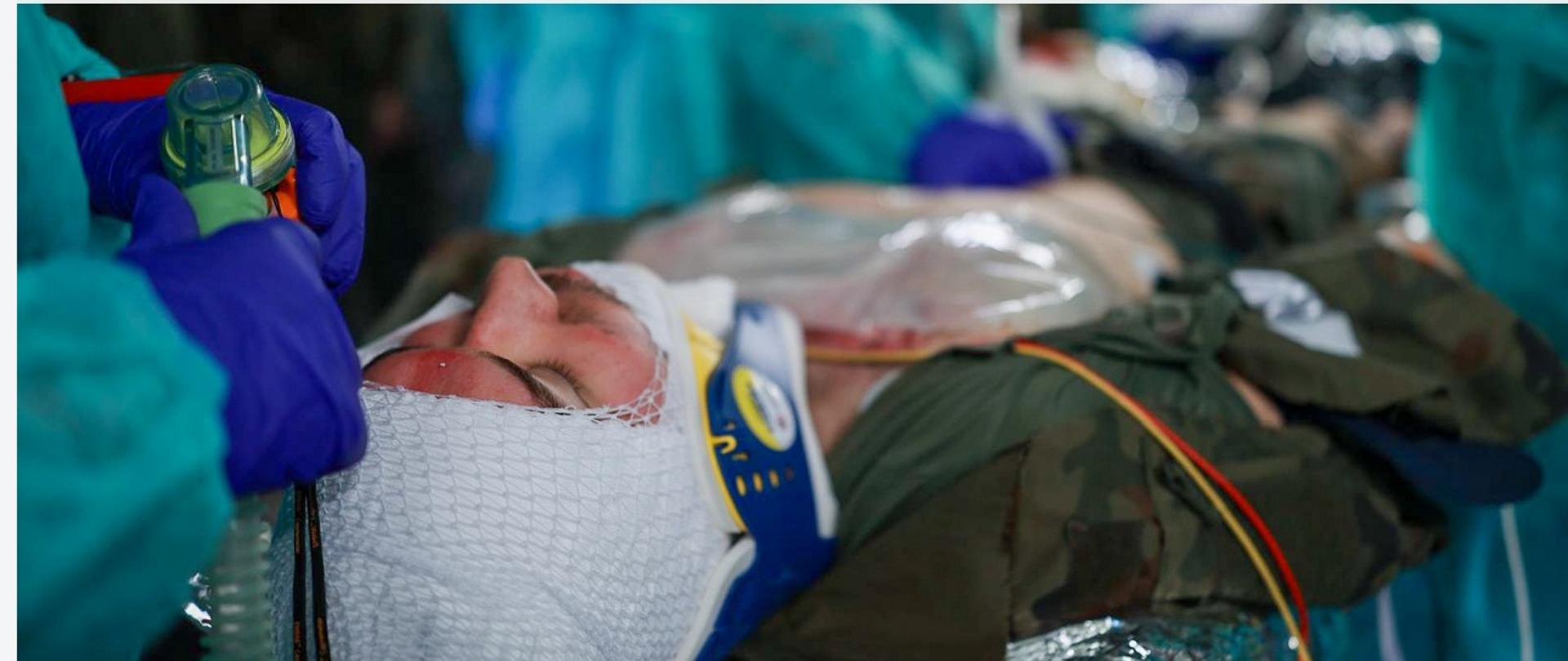 żołnierz z zabandażowana głowa leży na szpitalnym łóżku, wokół medycy