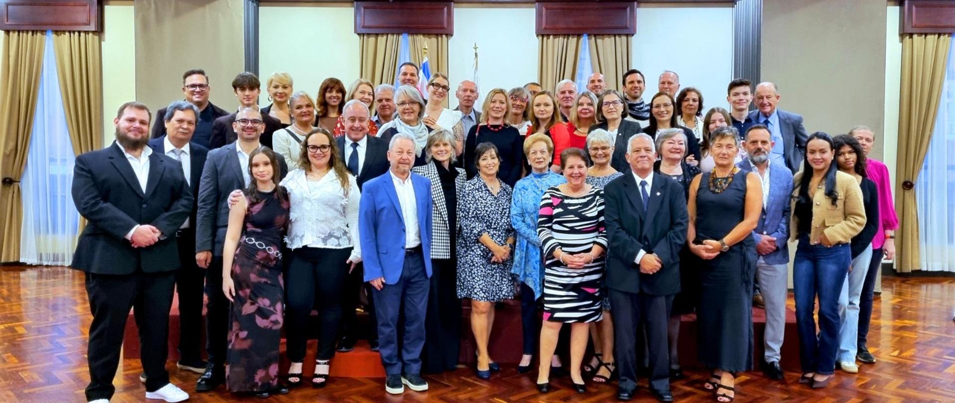 En la foto se ve a un grupo numeroso y elegantemente vestido, posando para una fotografía grupal en un salón claro y representativo.
Descripción detallada:
El grupo está formado por unas cuarenta personas, acomodadas en varias filas. Todos miran hacia la cámara y sonríen. La edad de los participantes es diversa. Muchas personas visten ropa formal: trajes, sacos, camisas, vestidos de cóctel y conjuntos elegantes. El ambiente es solemne.