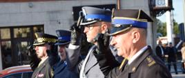 Strażak, policjanci i strażnik miejski stoją obok siebie i salutują. Wszyscy ubrani sa w mundury wyjściowe. W tle widać budynek i przejeżdżający samochód. 