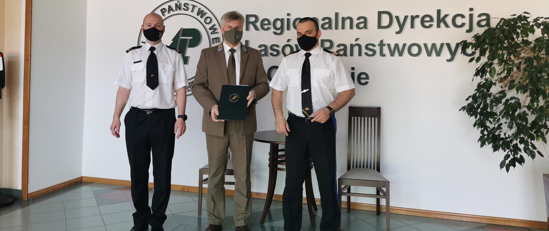 Komendant Wojewódzki PSP, Komendant Powiatowy PSP i Dyrektor Regionalnej Dyrekcji Lasów Państwowych w Olsztynie stoją na korytarzu obok siebie na tle RDLP. Dyrektor trzyma w ręku podpisane porozumienie. 