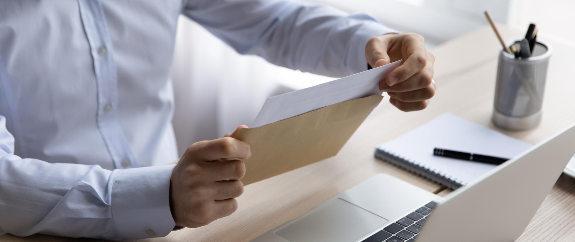 Mężczyzna w białej koszuli siedzący przed otwartym laptopem, w rękach trzyma papierową kopertę