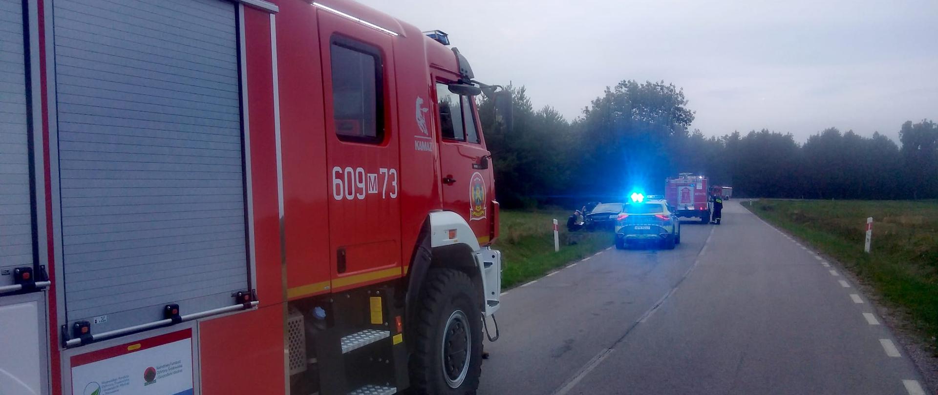 Zdjęcie przedstawia scenę z drogi, gdzie po lewej stronie znajduje się duży, czerwony wóz strażacki z numerem "609/73" i zamkniętymi żaluzjami na boku. Na tle wozu widoczne są różne naklejki informacyjne. Droga jest asfaltowa, a w tle widać inne pojazdy ratunkowe: policyjny radiowóz z włączonymi niebieskimi światłami oraz kolejny czerwony wóz strażacki. W oddali rozciąga się zielony krajobraz drzew i pól pod zachmurzonym niebem. Całość sugeruje interwencję służb ratunkowych, prawdopodobnie wypadek drogowy.