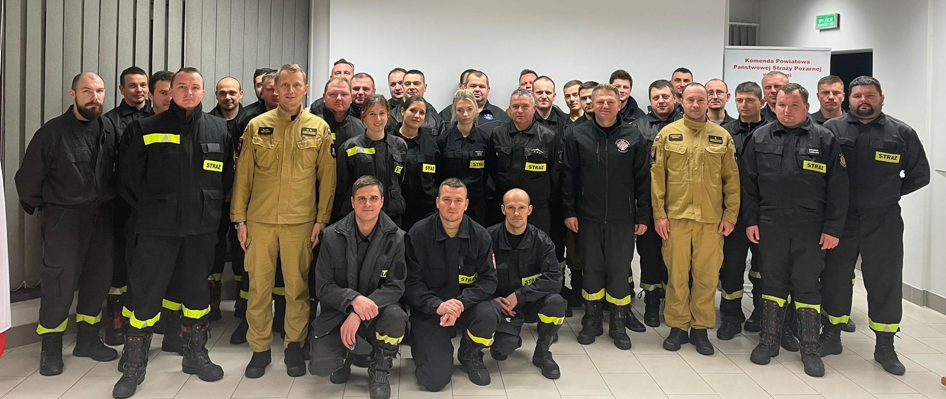 Kilkudziesięciu mężczyzn ubranych w czarne ubrania koszarowe strażackie z odblaskowymi paskami, czarnym butami stoją na Sali konferencyjnej. W tle ściany w kolorze białym, żaluzje koloru szarego