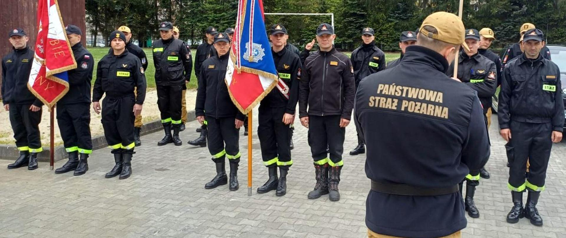 Zdjęcie zrobione na zewnątrz, na placu przed komendą podczas szkolenia członków pocztów sztandarowych. Strażacy stoją w szeregach, część z nich trzyma drzewce sztandarów.