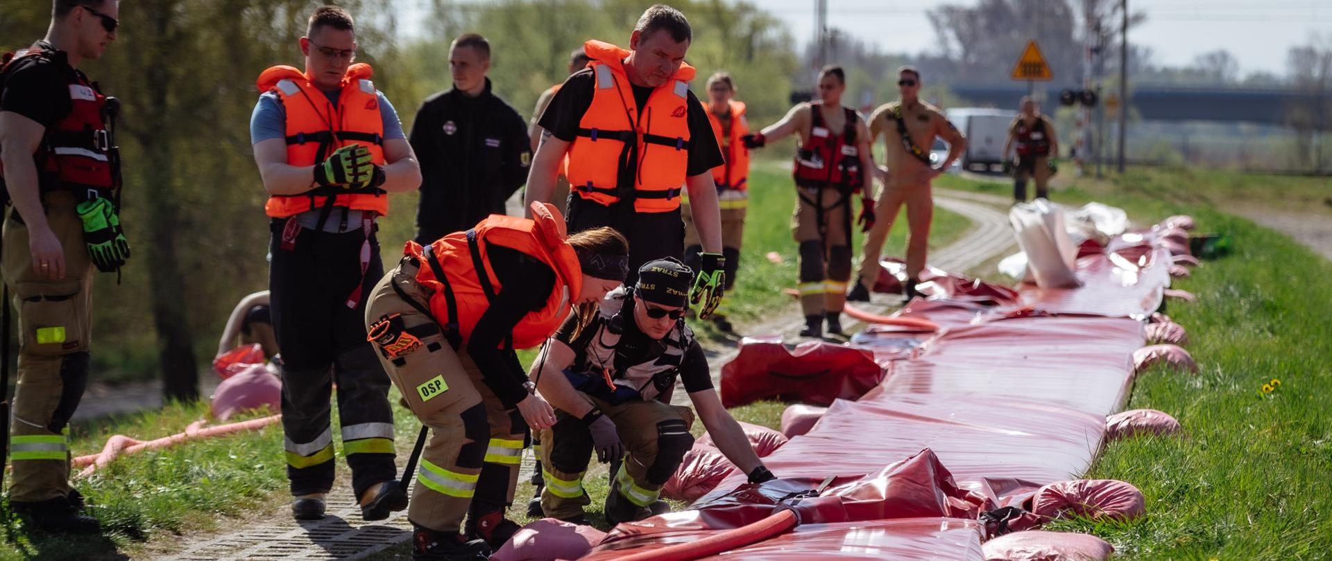 Strażacy w kamizelkach ratunkowych i ubraniach ochronnych układają i napełniają długi rękaw przeciwpowodziowy wzdłuż drogi. Na pierwszym planie dwie osoby regulują elementy instalacji, a w tle pozostali uczestnicy akcji przygotowują kolejne odcinki zabezpieczenia na terenie zielonym.