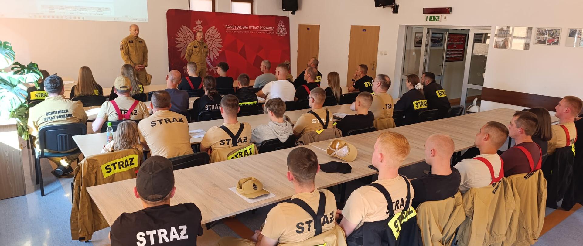 Zdjęcie przedstawia salę szkoleniową w KP PSP Sokołów Podlaski. Na pierwszym planie widzimy druhów osp w ubraniach koszarowych koloru piaskowego i czarnego siedzących za ławkami. Na drugim planie widzimy funkcjonariuszy PSP na tle czerwonego banera. 
