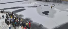 Fotografia przedstawia ujęcie wykonane z drona. Na fotografii widoczna tafla zamarzniętego jeziora, na brzegu stoi grupa uczniów szkoły podstawowej, na tafli lodu strażak pozoruje wpadnięcie do wody.