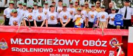 Zdjęcie zrobione na zewnątrz podczas uroczystej zbiórki uczestników obozu Młodzieżowych Drużyn Pożarniczych. W szeregu stoją ustawieni uczestniczy obozu oraz funkcjonariusze Państwowej Straży Pożarnej. Przed nimi ustawiony jest banner z napisem młodzieżowy obóz szkoleniowo-wypoczynkowy Solina 2024