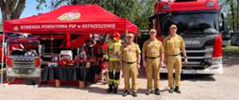 Stoisko Państwowej Straży Pożarnej z funkcjonariuszami stojącymi obok oraz samochodami strażackimi na drugim planie.