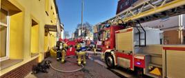  Ul. Piastowska, po prawej samochód pożarniczy drabina mechaniczna. W tle 2 karetki oraz samochód gaśniczy. 5 strażaków w mundurach bojowych i hełmach, trzech z aparatami oddechowymi. Do drzwi wejściowych doprowadzony wąż strażacki.