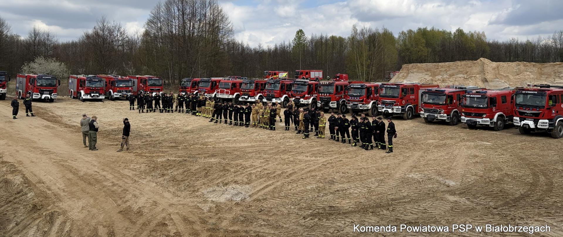 Pamiątkowe zdjęcie grupowe na piaszczystym poligonie. Kilkudziesięciu strażaków stoi w dwuszeregu przed długą linią kilkunastu czerwonych wozów strażackich różnych typów. W tle widać las i zachmurzone niebo.