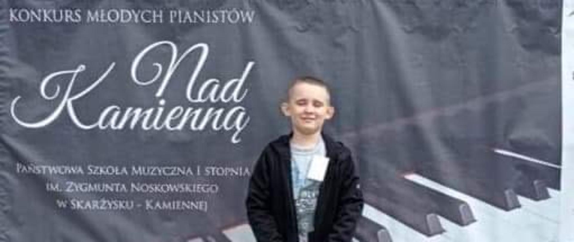 Chłopiec stoi na tle baneru z napisem Konkurs Młodych Pianistów "Nad Kamienną".