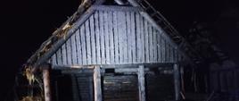 Zdjęcie w porze nocnej. Widać nadpalony budynek drewniany. Podłoże trawiaste. Oświetlenie sztuczne.