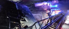 Widok z kosza drabiny mechanicznej, która znajduje się nad dachem budynku w którym doszło do pożaru. W tle strażacy prowadzący dogaszanie.