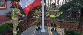 Przy maszcie flagowym poczet flagowy strażaków ubranych w ubrania specjalne i hełmy koloru czerwonego. Z lewej strony strażak przypina Flagę Polski do masztu. W tle drzewa. Dzień jest słoneczny.