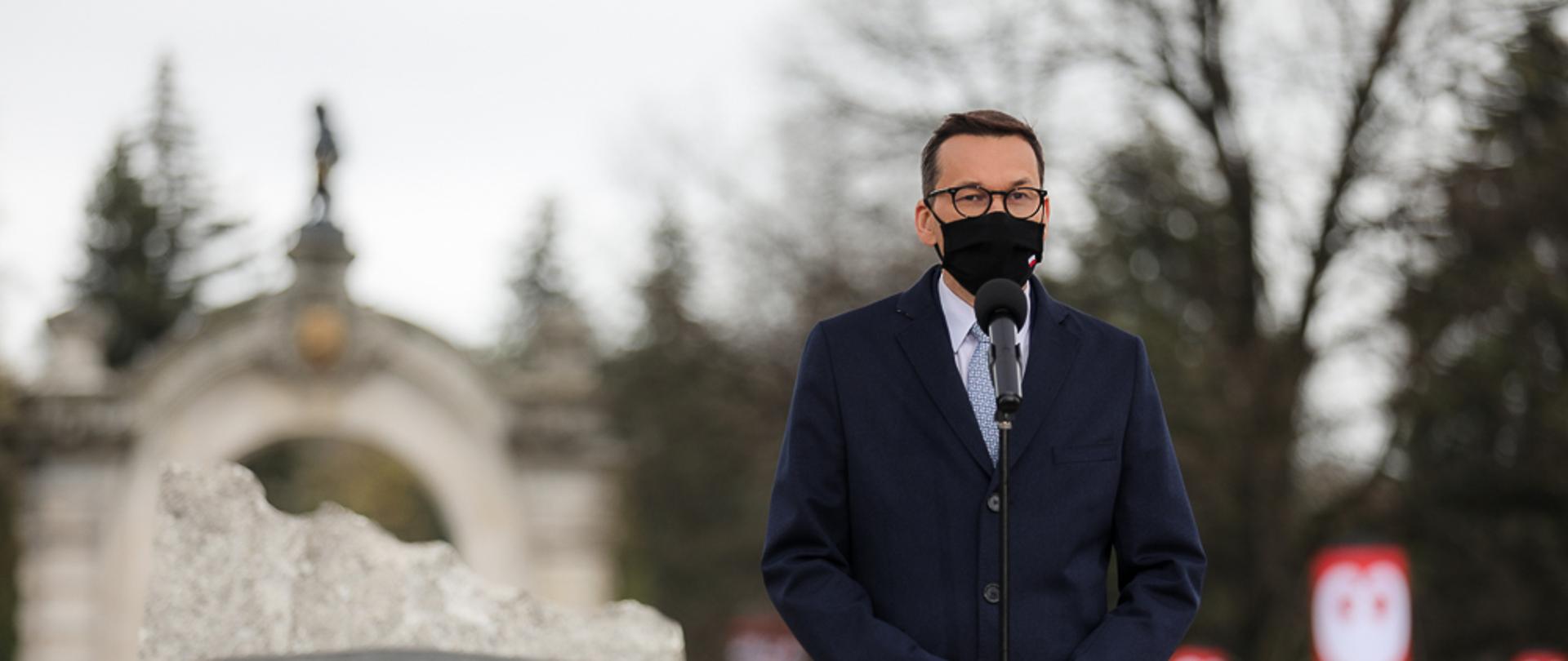 Premier Mateusz Morawiecki stoi przy mikrofonie w tle pomnik.
