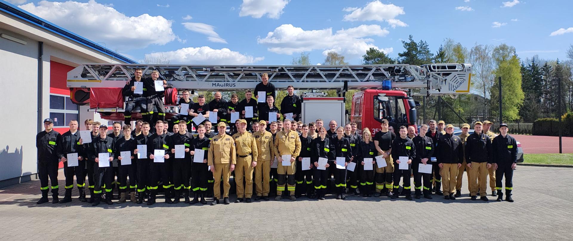 Grupa kilkudziesięciu strażaków w umundurowaniu stoi przed remizą strażacką i samochodem z drabiną mechaniczną Magirus. Część osób trzyma w rękach dokumenty lub certyfikaty. Zdjęcie wykonane na zewnątrz w słoneczny dzień, na tle nieba z chmurami.