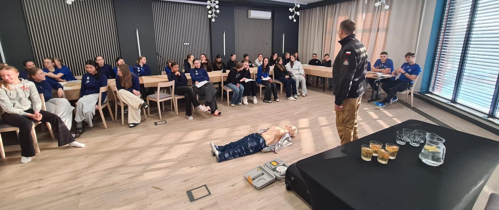 Zdjęcie dokumentuje szkolenie z zakresu pierwszej pomocy, podczas którego instruktor prowadzi zajęcia dla grupy młodzieży w nowoczesnej sali konferencyjnej. Na pierwszym planie widoczny jest manekin do resuscytacji oraz defibrylator AED, służące do praktycznej nauki ratowania życia.