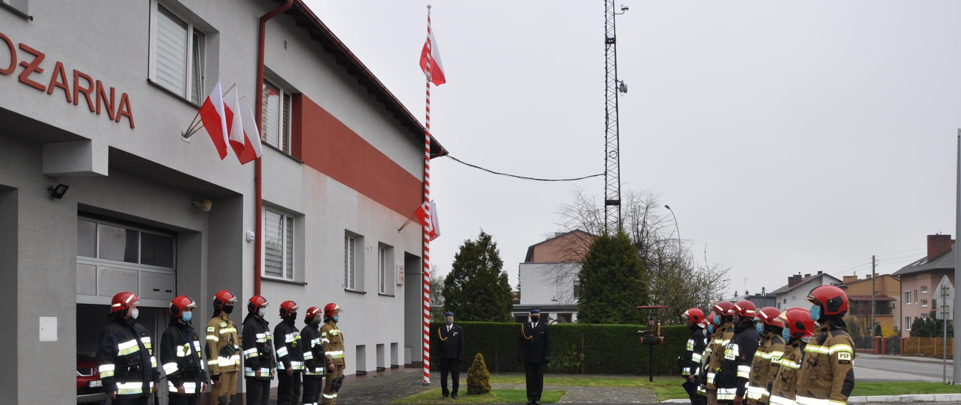 Na zdjęciu widzimy na pierwszym planie strażaków w pełnym uzbrojeniu, którzy dokonują podniesienia Flagi państwowej na maszt. Na drugim planie widać pozostałych strażaków stojących w szeregu oraz komendanta. W tle widać budynek komendy. 