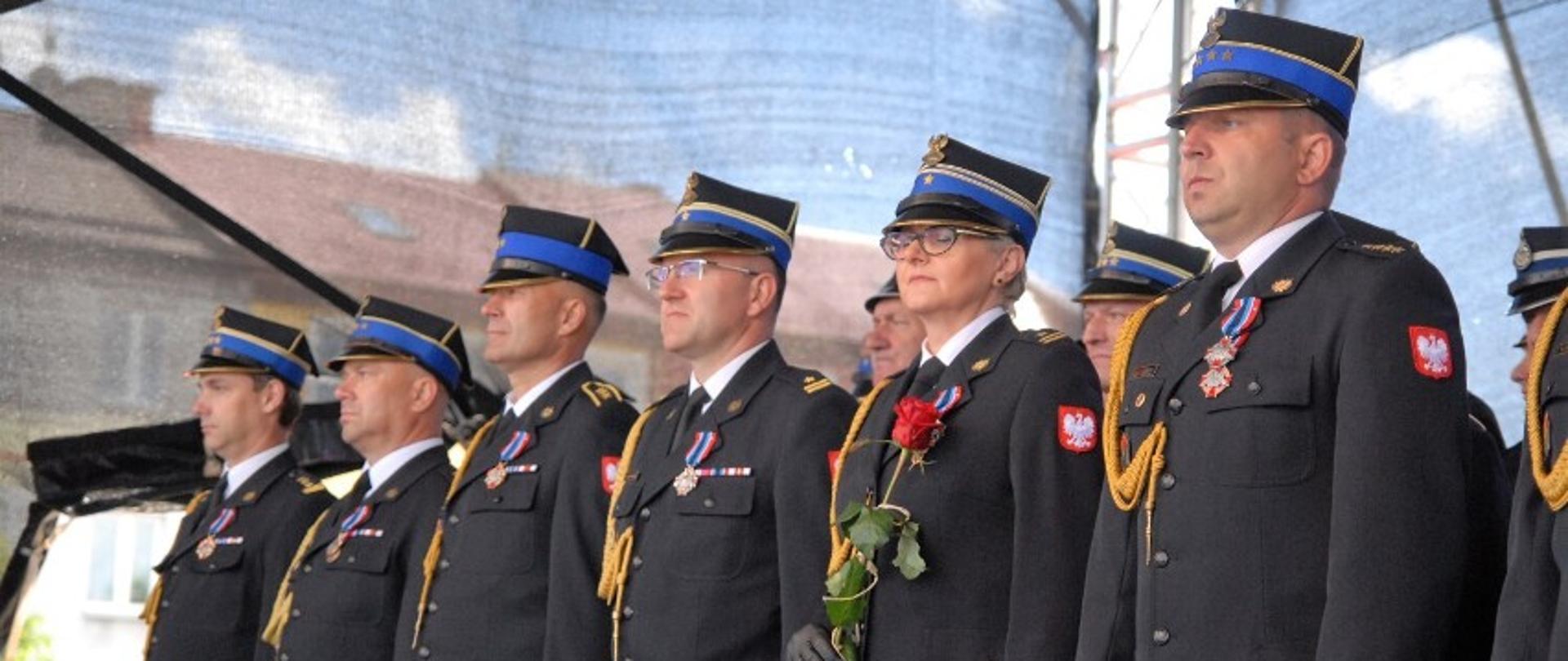 Zdjęcie przedstawia sześciu funkcjonariuszy Państwowej Straży Pożarnej w umundurowaniach wyjściowych odznaczonych odznakami Zasłużony dla Ochrony Przeciwpożarowej. Jeden z funkcjonariuszy (kobieta) trzyma w ręku czerwoną różę