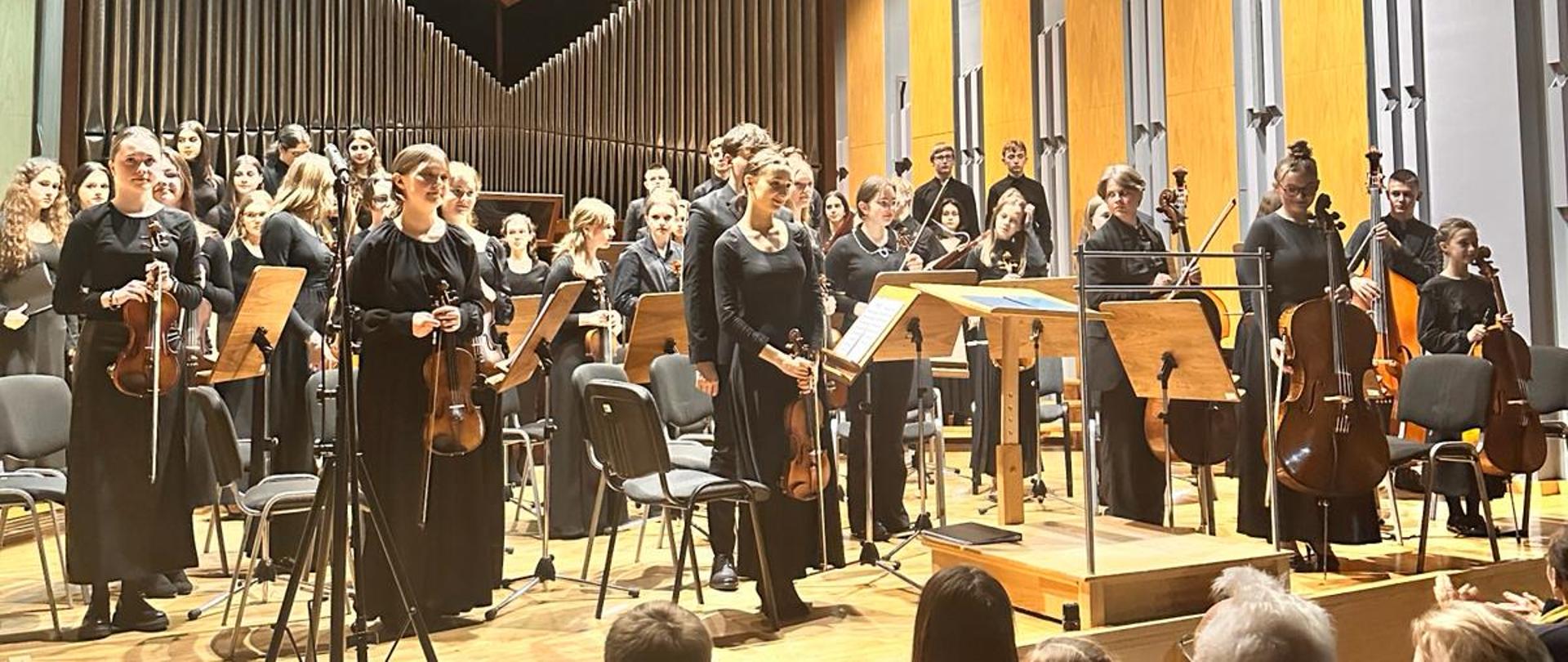 Zdjęcie na sali koncertowej. Na pierwszym planie publiczność siedząca na widowni. Na scenie sali koncertowej wykonawcy orkiestry stoją trzymając w dłoni swoje instrumenty. Za wykonawcami puste krzesła, przed nimi pulpity. W tle organy.