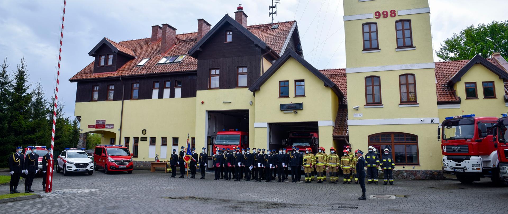 Uroczysta zbiórka na palcu komendy. NA placu stoją strażacy, naprzeciwko nich stoi dowódca uroczystości, który oddaje honor podczas podnoszenia flagi państwowej na maszt. Za strażakami znajduje się budynek komendy i widoczne są samochody strażackie. 