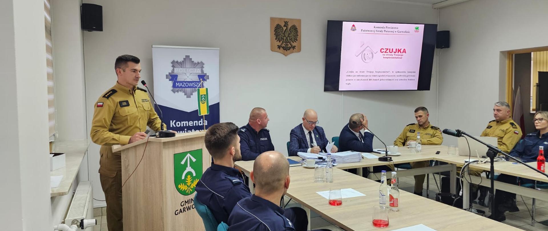 Prelegent w mundurze służbowym stoi przy mównicy, prezentując slajd na ekranie z napisem "CZUJKA na straży Twojego bezpieczeństwa" i logo Komendy Powiatowej Policji w Garwolinie. Przy stole konferencyjnym w głębi siedzi grupa osób w mundurach policyjnych i cywilnych, skupionych na dokumentach i prezentacji.