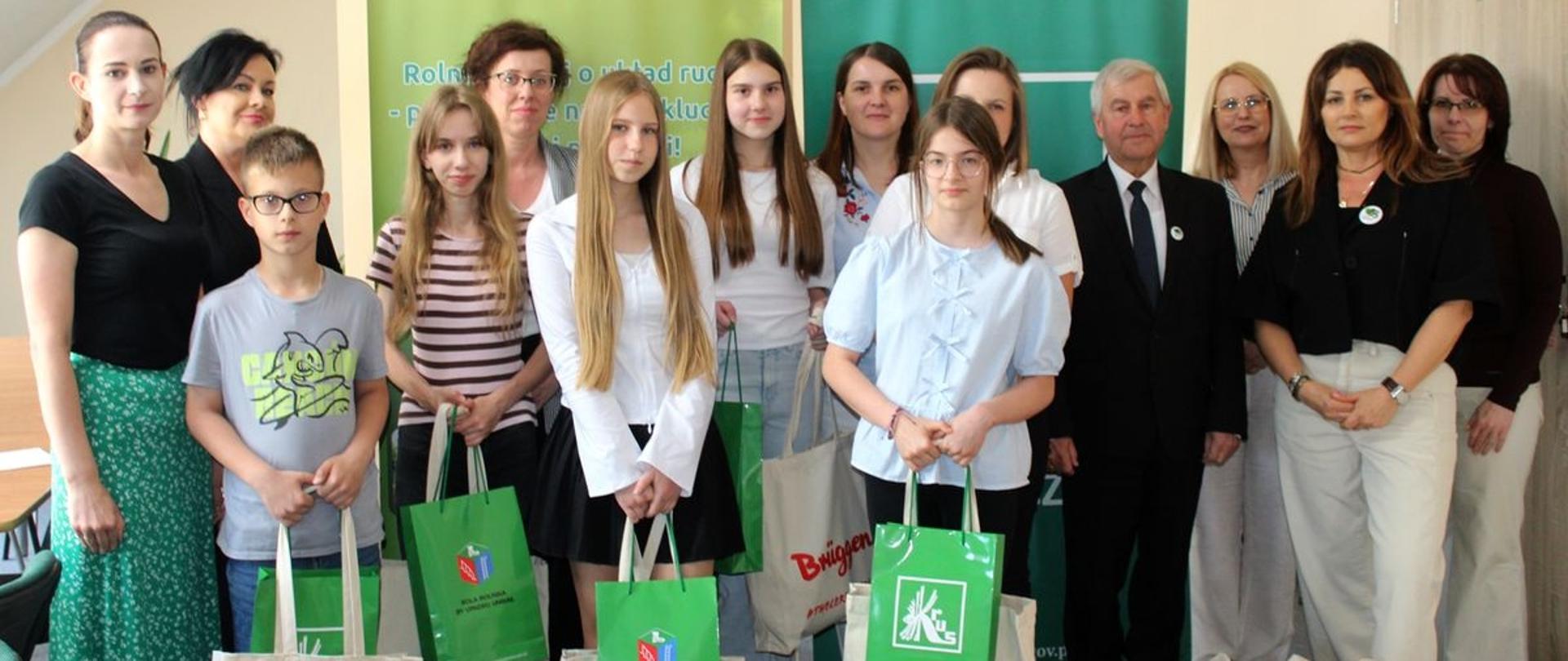W sali urzędu na tle dwóch roll-upów z logo KRUS pozują do zdjęcia mężczyzna, 8 kobiet oraz 5 dzieci (4 dziewczynki i 1 chłopiec). Każde dziecko trzyma w ręce 2 torby z nagrodami. Wszyscy uśmiechają się do zdjęcia.