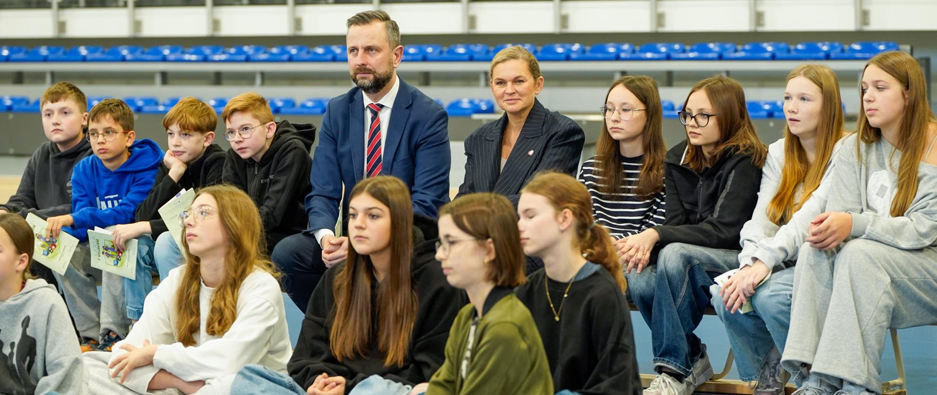 Minister Edukacji Barbara Nowacka i mężczyzna w garniturze siedzą na krzesłach, otoczeni dużą grupą młodzieży siedzącej na ławkach i na niebieskiej podłodze hali sportowej, obserwując wydarzenie.