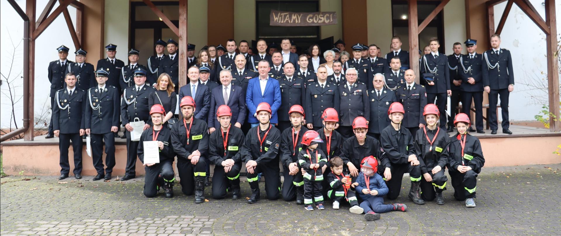 Druhowie OSP strażacy PSP przedstawiciele władzy rządowej i samorządowej oraz młodzież z młodzieżowej drużyny pożarniczej pozują do zdjęcia na tle budynku remizy osp