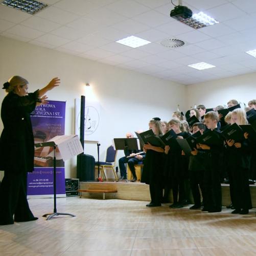 Na scenie stoi chór ubrany na czarno. Przed chórem stoi kobieta dyrygent również ubrana na czarno. 