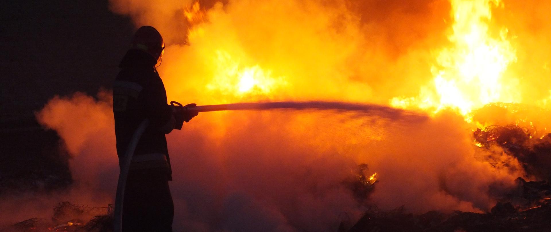 Strażak gasi pożar. W tle pomarańczowe płomienie i kłęby szarego dymu. 