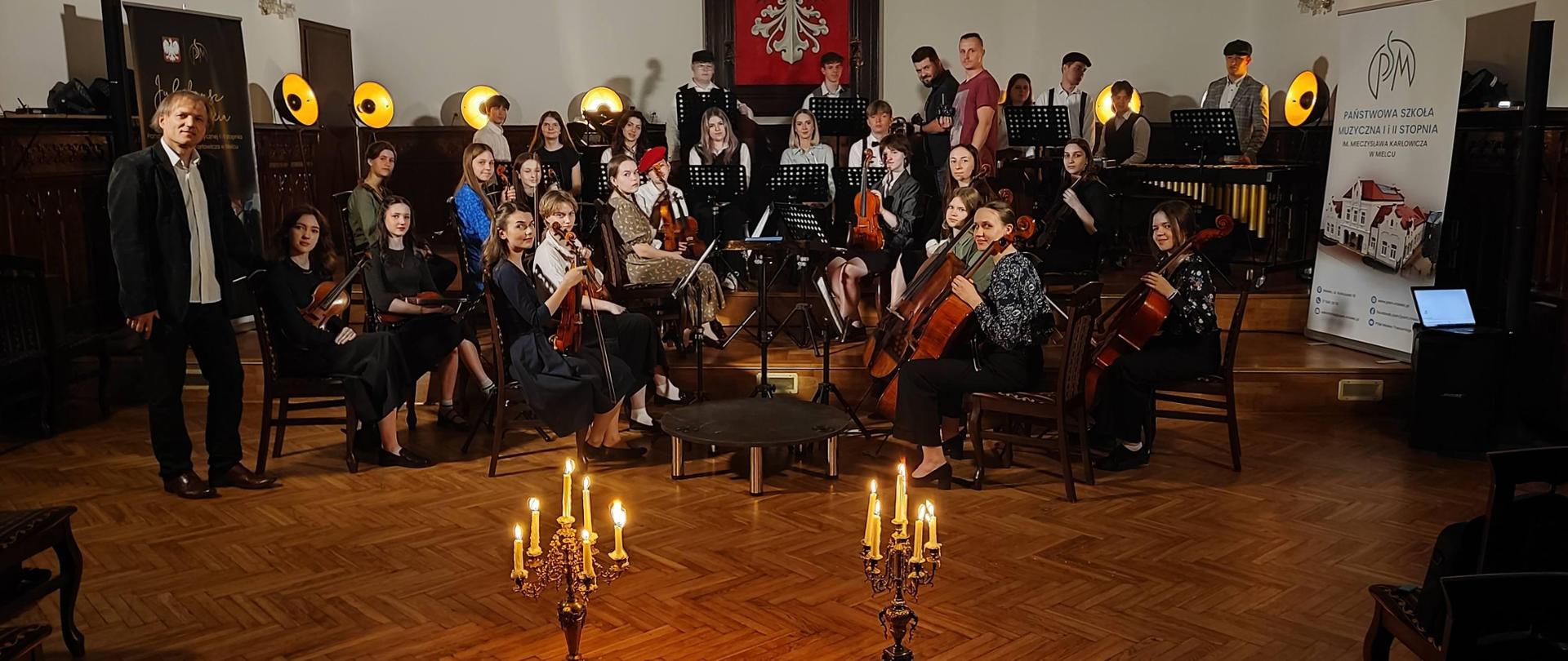 Orkiestra PSM Mielec na klimatycznym zdjęciu, z dwoma świecznikami z przodu i ciepłym osiwetleniem scenicznym w tle na scenie w sali królewskiej PSM Mielec