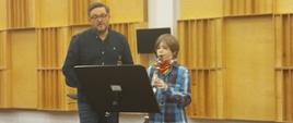 Młody uczeń gra na klarnecie, a nauczyciel daje mu wskazówki gry, w tle widoczna jest klasa muzyczna