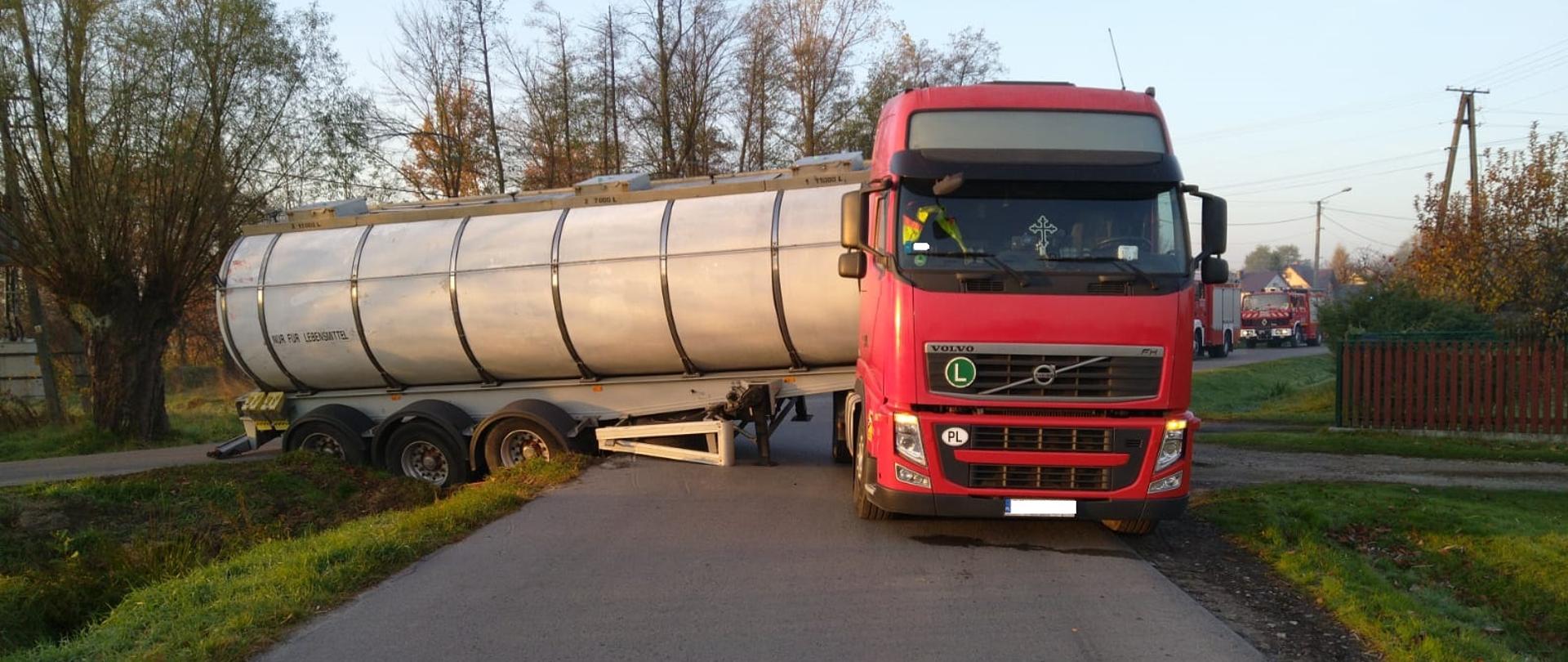 Ciągnik siodłowy koloru czerwonego z cysterna na drodze, prawe koła cysterny na bocznej drodze osunięte na rów drogi, naczepa cysterny mocno przechylona, droga zablokowana, w oddali widać pojazdy Państwowej i Ochotnicznej Straży Pożarnej.
