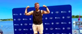 Zawodnik stoi na podium z uniesionymi rękoma, na tle niebieskiego baneru Ministerstwa Sportu i Turystyki